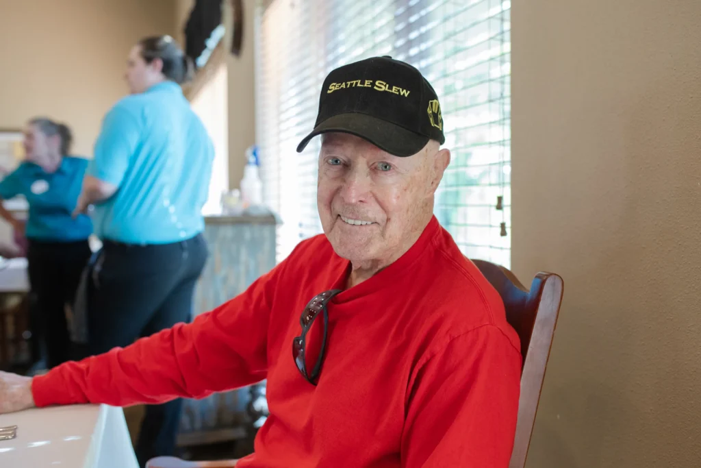 A community resident man is sitting at a table, looking directly at the camera and smiling. He is wearing a black cap, and a red shirt.