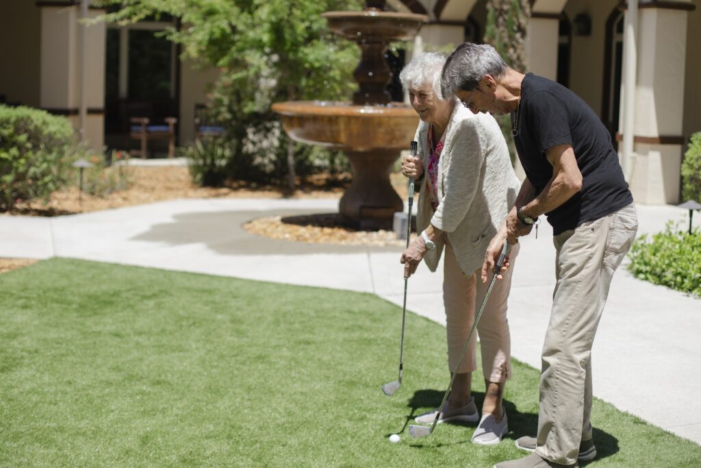 An elderly woman and man are putting on a small grassy area with a fountain in the background. Both are holding a golf club. She is smiling.