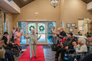 A community resident dressed in a light-colored suit walks down a red carpet with arms outstretched, welcomed by a group of residents and caregivers clapping and cheering in a warmly decorated community space