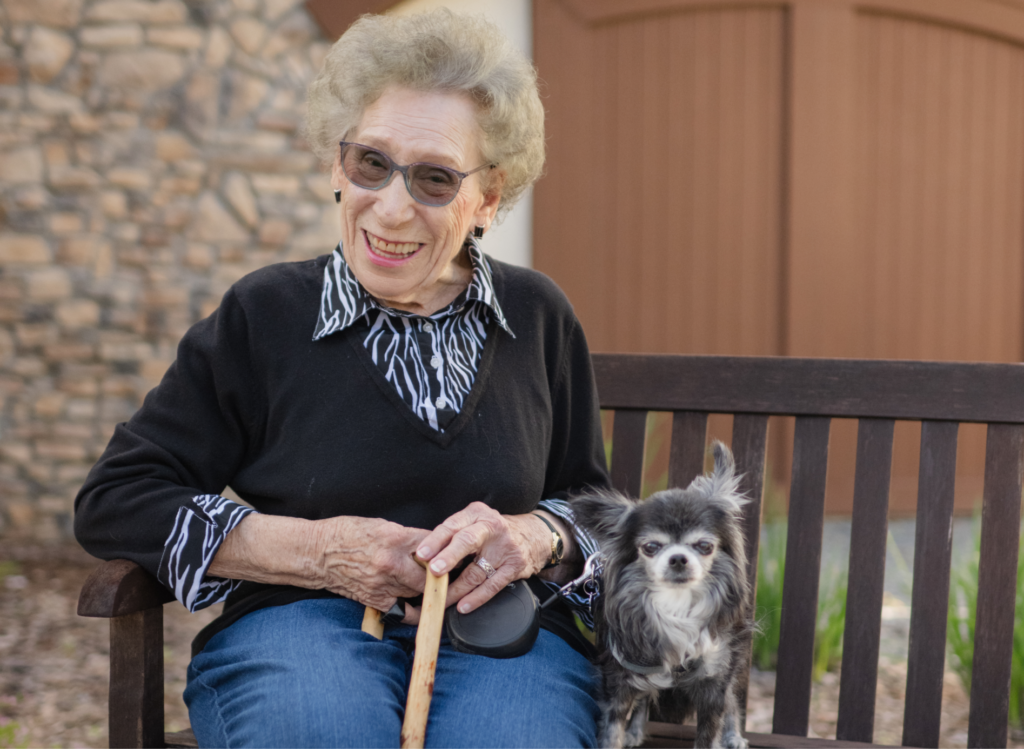 A community resident with glasses, smiling and holding a cane, sits on a wooden bench next to a small dog. The woman wears a black sweater over a striped shirt, and the dog is sitting calmly beside her. A stone wall and wooden doors are in the background.