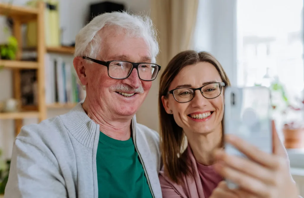 Smiling community resident with glasses and white hair standing next to a younger woman with glasses, both talking to someone on the smartphone in a cozy, well-lit home setting