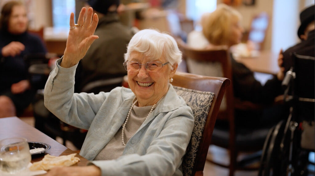 Smiling community resident waving while seated at a dining table. Other residents are visible in the warmly lit background, enjoying their meals and conversations