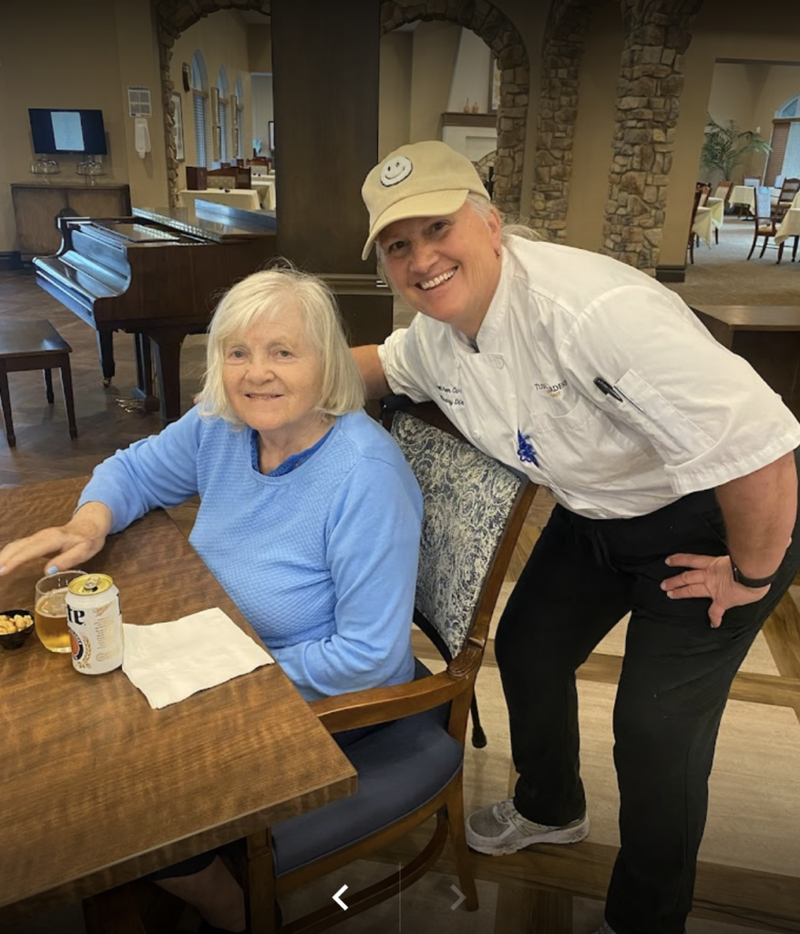 An experienced Tuscan Garden's associate poses next to a community resident who's sitting at a table. Both smile while looking at the camera.