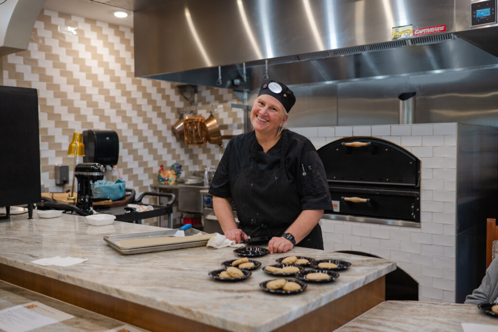 A smiling chef in a black uniform and hat stands behind a marble counter in a modern kitchen. Various pastries on plates are in the foreground. A brick oven and kitchen equipment are visible in the background.