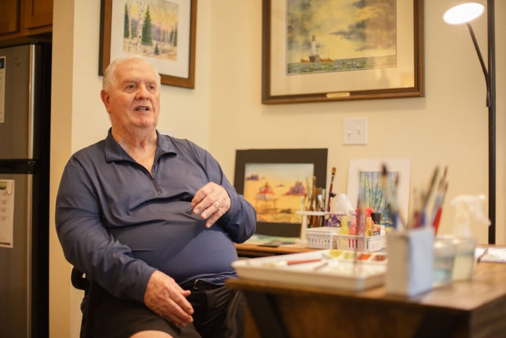 Community resident sitting in a cozy living space, engaged in conversation with a relaxed expression. A table with various items and framed artwork on the wall create a warm atmosphere