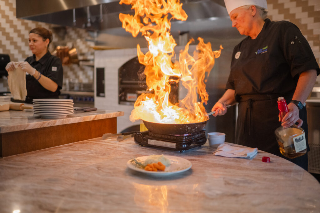 A chef in a white hat and black AgeWell uniform flambéing a dish in a pan with large flames, holding a bottle of alcohol. Another person in the background prepares food beside stacked plates. A plate with food is on the counter in the foreground.