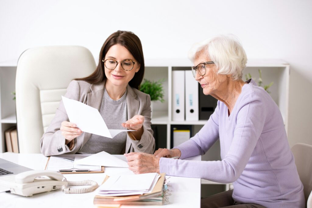 A woman with glasses and a blazer sits at a desk, engages in a financial planning conversation with a community resident wearing glasses and a lavender sweater.