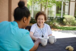 An older woman smiling and enjoying coffee outdoors with a team member at a senior living community.