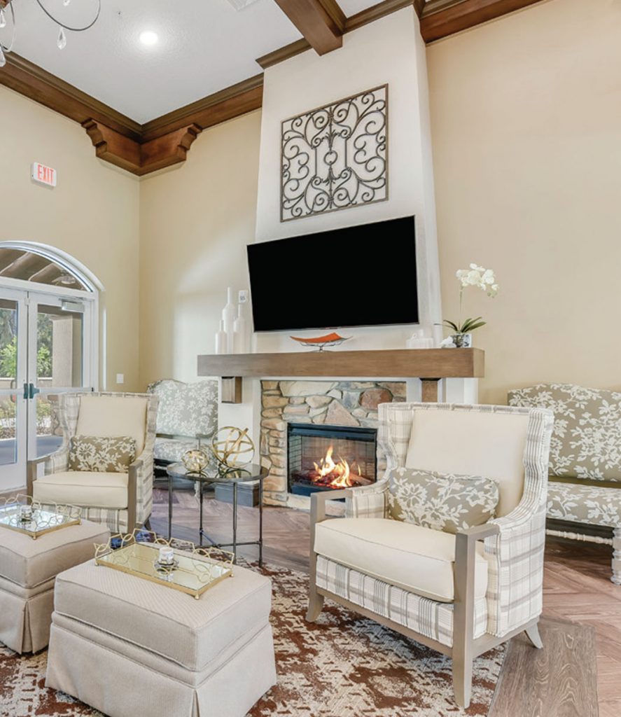 Elegant living room with tall ceilings, featuring a fireplace below a wall-mounted TV. Cream-colored chairs and a floral-patterned sofa surround a glass coffee table. Decorative vases and a firewood nook complete the cozy ambiance.