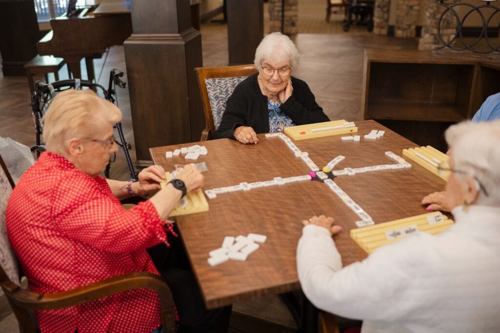 Four community residents sit around a square table playing a game of dominoes. They are focused on the game, with domino tiles arranged in front of them. The room has a warm, wooden interior.